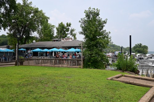 A lakeside restaurant with outdoor seating, surrounded by trees and boats, under a cloudy sky.