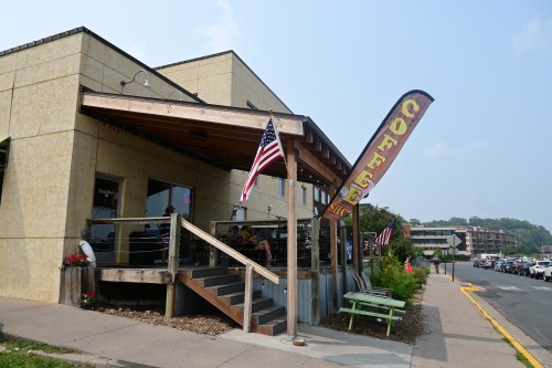 A coffee shop with a wooden deck, American flags, and outdoor seating on a sunny day.