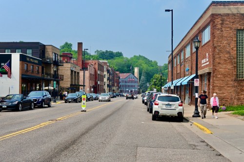 A street scene with parked cars, shops, and pedestrians on a sunny day, surrounded by green hills in the background.