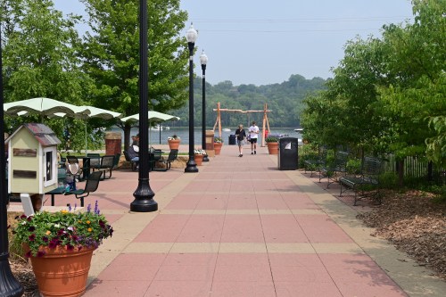A scenic riverside walkway with benches, planters, and people strolling under green trees on a sunny day.