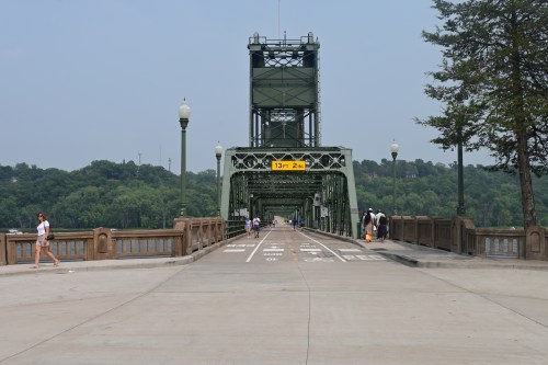A green bridge with a height warning sign, surrounded by trees and people walking along the pathway.