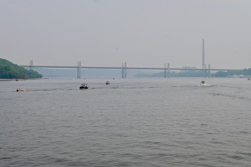 A river scene with boats navigating under a bridge, surrounded by hazy skies and green hills in the background.