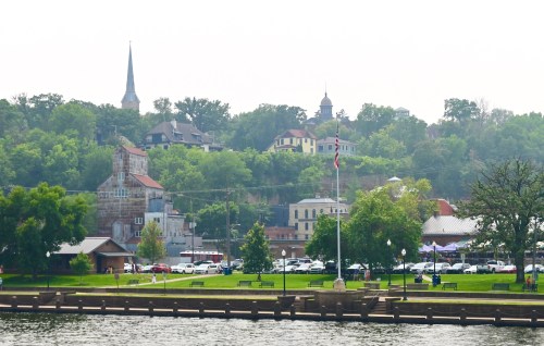 A scenic view of a riverside town with green hills, historic buildings, and a church steeple in the background.