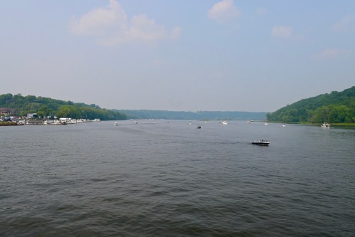A serene river scene with boats on the water, surrounded by lush green hills under a blue sky with scattered clouds.