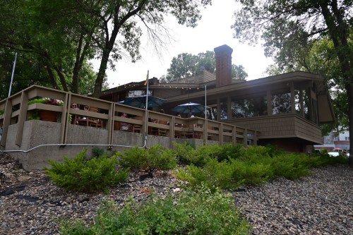 A modern restaurant with outdoor seating, surrounded by trees and greenery, featuring umbrellas and a stone pathway.