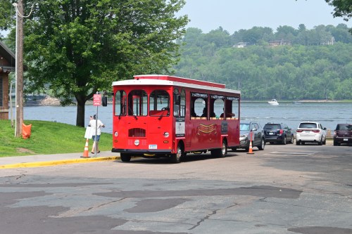 A red trolley bus parked by a river, surrounded by trees and cars on a sunny day.