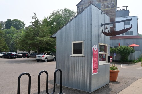 A small, silver metal kiosk with a sign, surrounded by trees and parked cars in a lot.