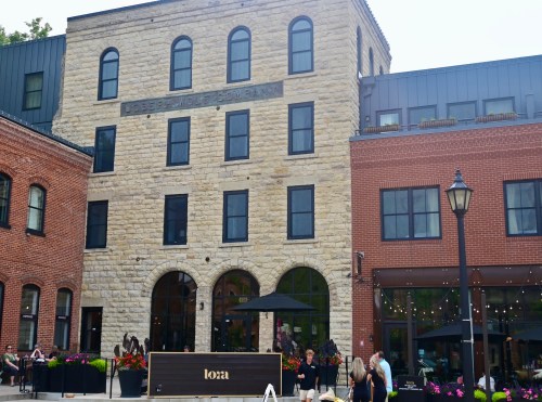 Historic stone building with modern additions, outdoor seating, and vibrant flowers in a lively urban setting.