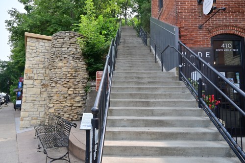 Concrete stairs lead up from a sidewalk, flanked by a stone wall and a brick building with a sign. Lush greenery surrounds.