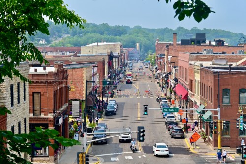 A bustling street scene with shops, cars, and pedestrians, framed by trees and a clear sky in the background.