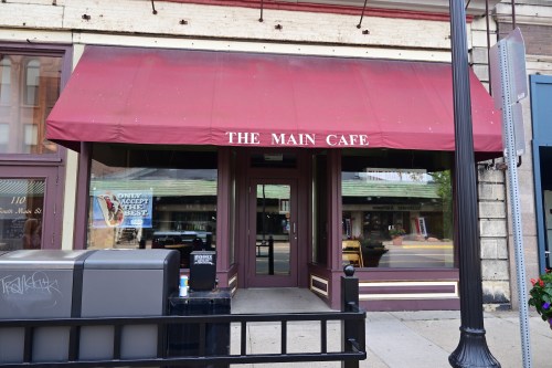 Exterior of The Main Cafe featuring a red awning and large windows, with a street view and nearby lamppost.
