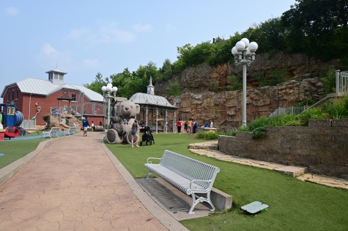 A park scene featuring a large teddy bear statue, playground equipment, and people enjoying the outdoors near rocky terrain.