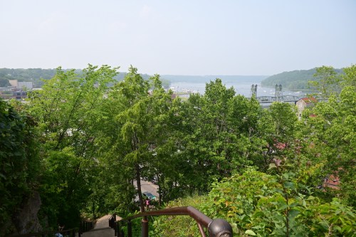 A scenic view from a hilltop, featuring lush green trees and a river with boats in the distance under a clear sky.