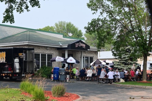 Outdoor dining scene at a restaurant with patrons seated at picnic tables under umbrellas.