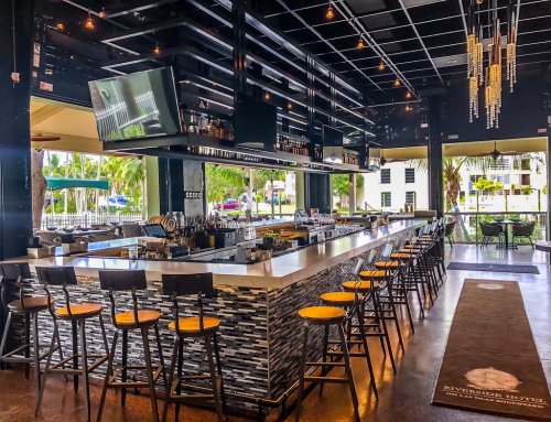Modern bar interior with a sleek design, high stools, and a large TV, surrounded by large windows and tropical views.