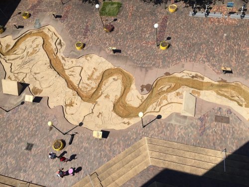 Aerial view of a winding sand river design on a plaza, with people walking nearby and planters around the edges.