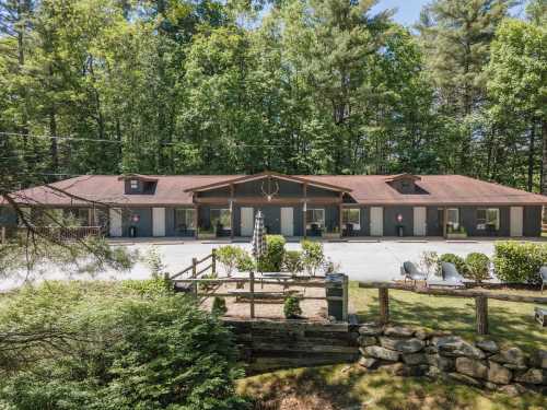 A rustic building surrounded by trees, featuring a gravel path, garden, and outdoor seating area.