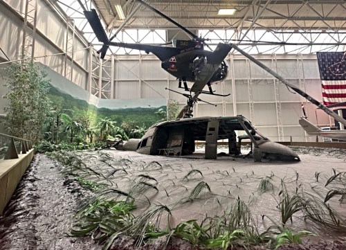 A military helicopter suspended above a damaged aircraft in a museum, surrounded by greenery and a mural of a jungle.