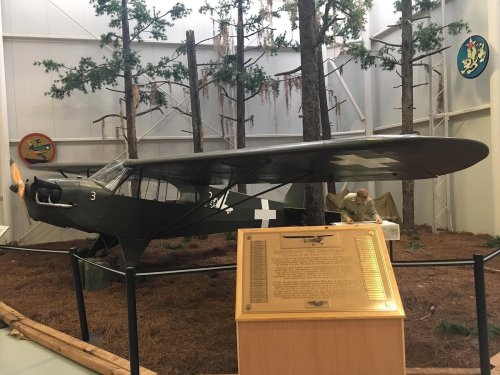 A vintage military aircraft displayed indoors among trees, with an informational plaque in front.