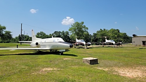 A small white aircraft in the foreground with several other planes parked on a grassy field under a clear blue sky.