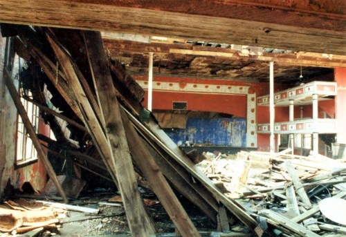 Interior of a dilapidated building with collapsed wooden beams and debris scattered on the floor.