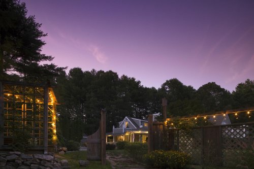 A cozy house at dusk, surrounded by trees and illuminated by string lights, creating a warm, inviting atmosphere.