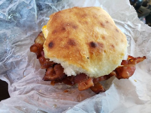 A close-up of a bacon sandwich on a toasted biscuit, wrapped in paper, with crispy bacon and a golden crust.
