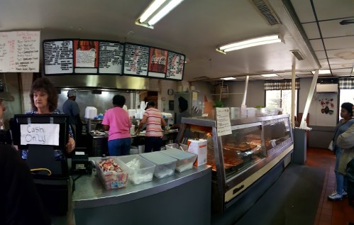 A busy diner interior with a counter, menu board, and customers ordering food. Cash-only sign visible.