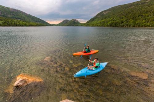 Two kayakers in colorful kayaks paddle on a calm lake surrounded by green hills under a cloudy sky.