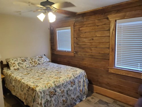 Cozy bedroom with a wooden accent wall, a bed with a floral quilt, and two windows with blinds.