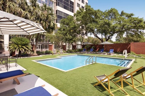 A sunny pool area with lounge chairs, umbrellas, and greenery surrounding a clear blue pool.