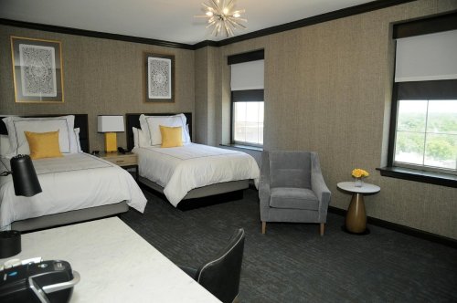 A modern hotel room with two beds, a gray chair, and large windows, featuring stylish decor and a small table.