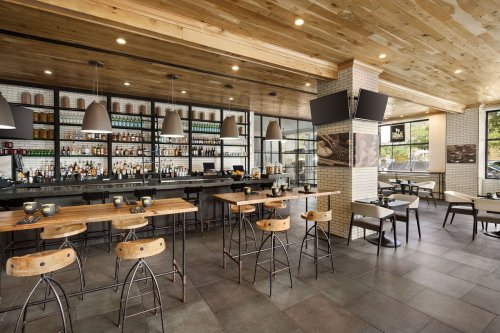 Modern restaurant interior featuring a wooden bar, high stools, and a well-stocked shelf of drinks. Bright and inviting atmosphere.