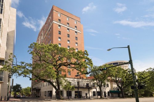 A tall brick building stands at the corner of a street, surrounded by trees and under a clear blue sky.