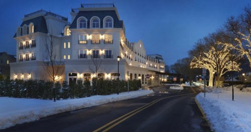 A charming hotel illuminated at dusk, surrounded by snow and lined with trees adorned with warm lights.