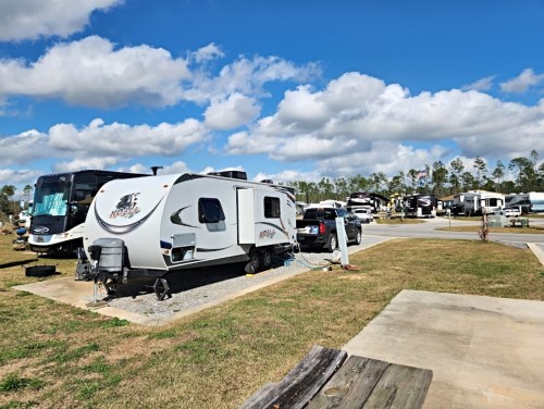 A parked RV in a campground with other RVs in the background under a blue sky with fluffy clouds.