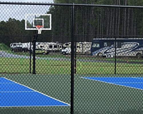 A basketball hoop in the foreground with blue courts, and RVs parked behind a chain-link fence in a wooded area.