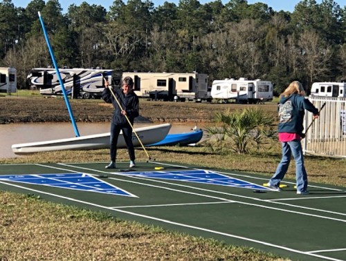 Two people playing shuffleboard on a green court near a body of water, with RVs parked in the background.