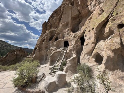 A rocky cliff with cave-like openings and a wooden ladder leading up to one of the entrances, under a cloudy sky.