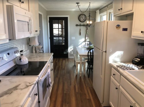 Bright kitchen with white cabinets, marble countertops, and a dining table by the door, featuring wooden floors and natural light.