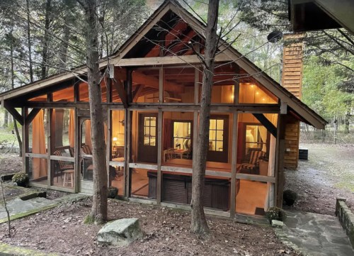 A cozy cabin with large windows, surrounded by trees, showcasing warm interior lighting and rustic furniture.
