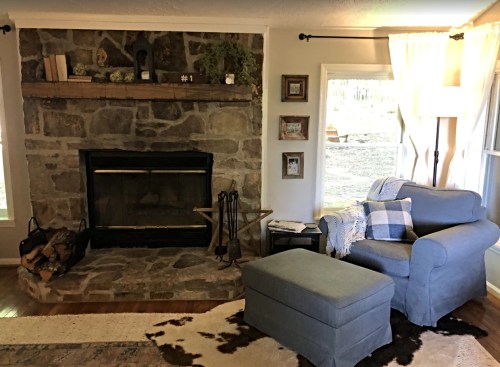Cozy living room with a stone fireplace, blue armchair, ottoman, and natural light from large windows.