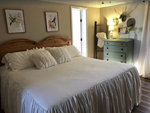 Cozy bedroom featuring a large bed with white bedding, a green dresser, and floral wall art. Natural light from a door.