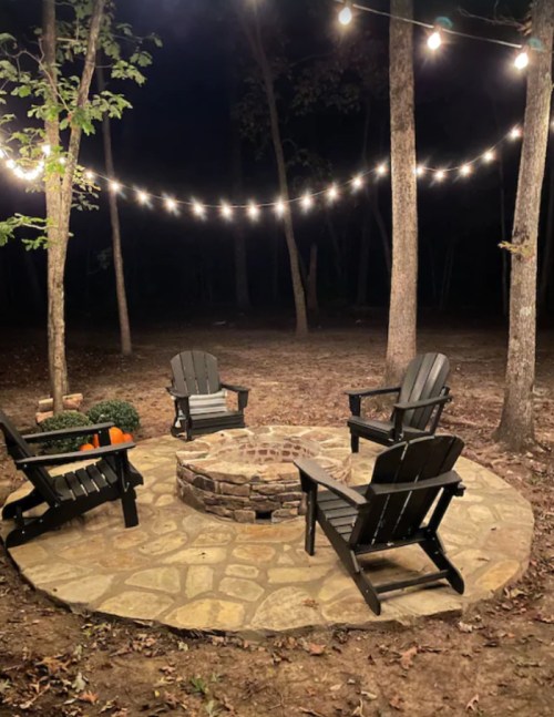 A cozy outdoor fire pit area with four black chairs, surrounded by trees and string lights at night.