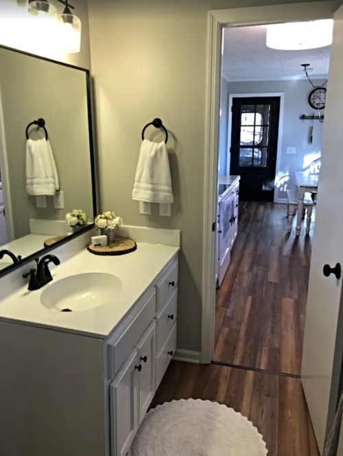 A modern bathroom with a white sink, mirror, and towels, leading to a kitchen area with wooden floors.