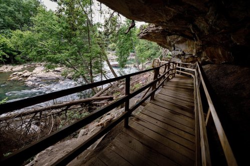 A wooden walkway leads through a rocky area beside a river, surrounded by lush green trees.