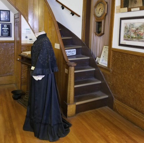 A vintage black dress on a mannequin beside a wooden staircase in a historic building.