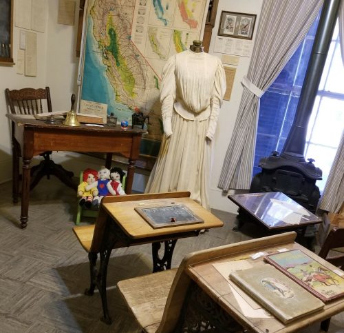 A vintage classroom scene featuring a dress form, old desks, a map, and toys, evoking a historical educational setting.