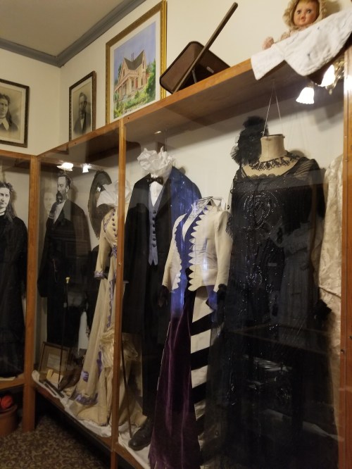 Display of vintage clothing and accessories in a museum, featuring formal wear and historical photographs.
