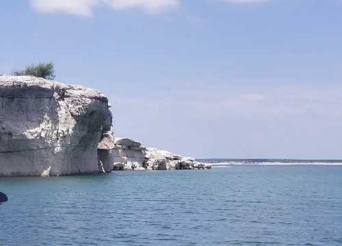 A rocky shoreline with cliffs beside calm blue water under a clear sky.